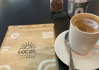 Image of a Local Coffee + Shop menu on a table next to a white coffee cup and saucer filled with a coffee drink.