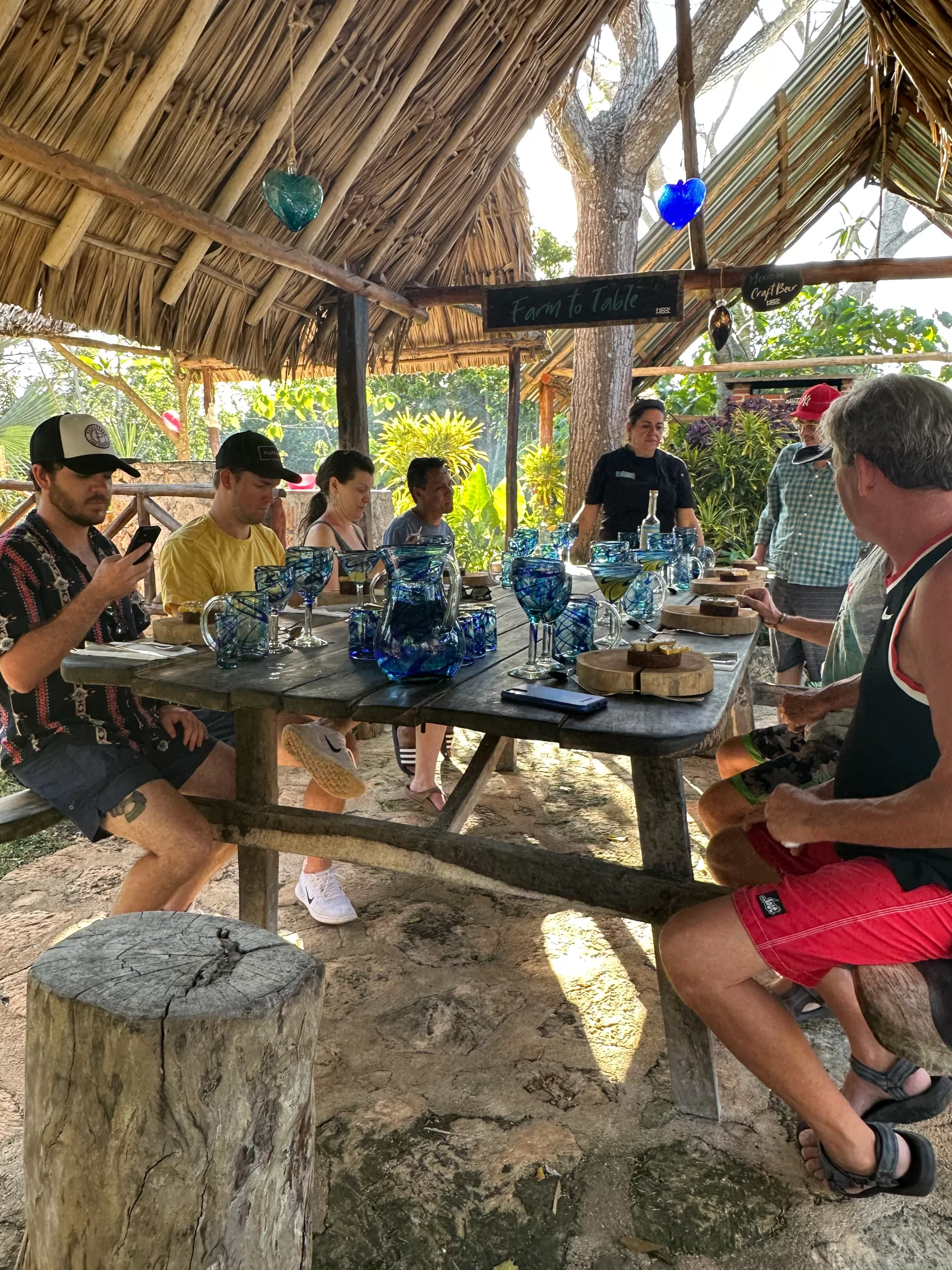 A group of five visitors sitting at a picnic table filled with blue glasswear and speaking with Chef Karla Romo.