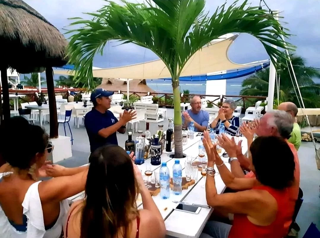 A group of people sitting around a white picnic table on the roof of La Sirena restaurant, enjoying Tequila University with an ocean view in Puerto Morelos.