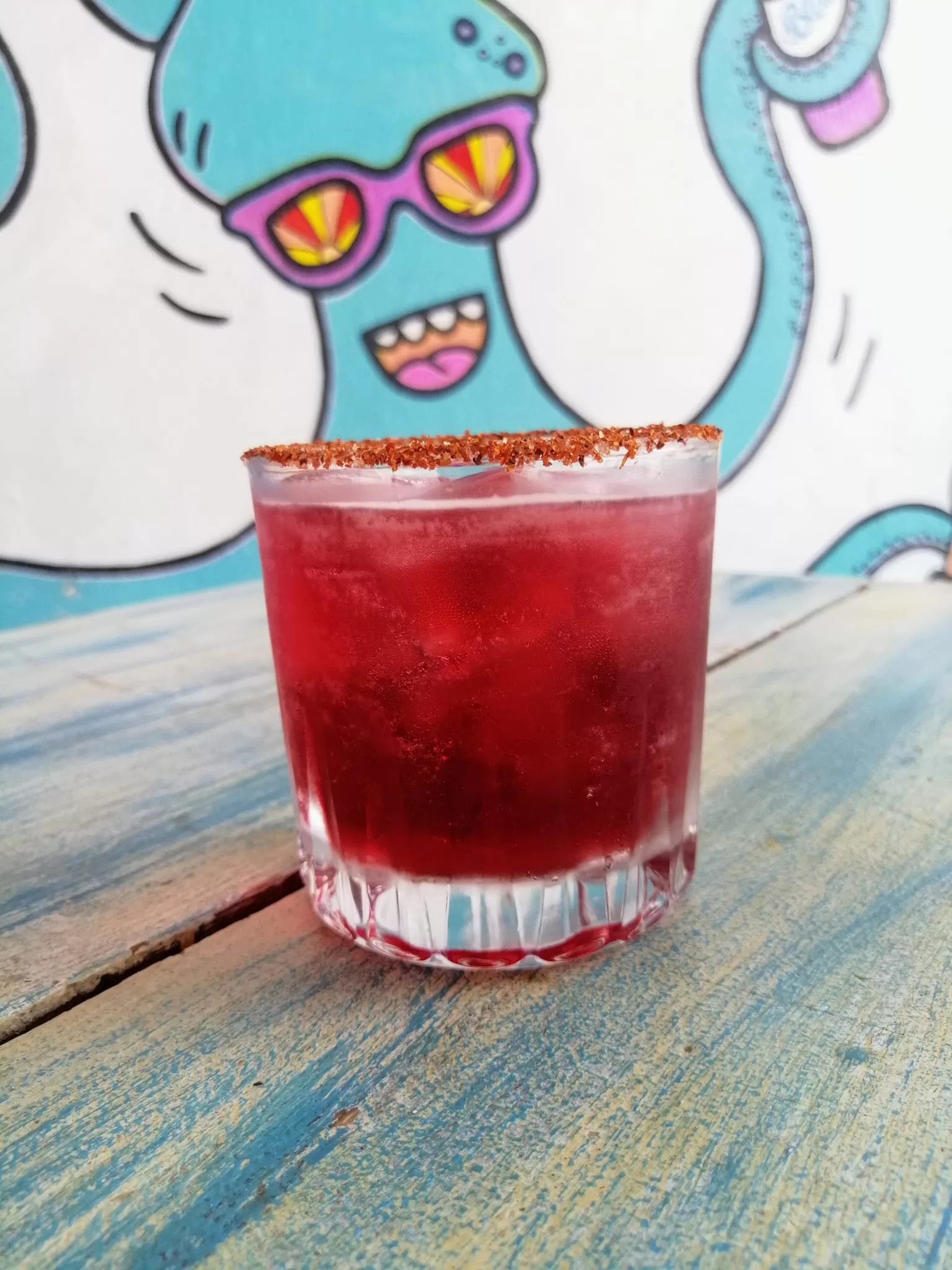 Glass rimmed with tajin and filled with red jamaica margarita. Blue octopus painted on the wall in the background.