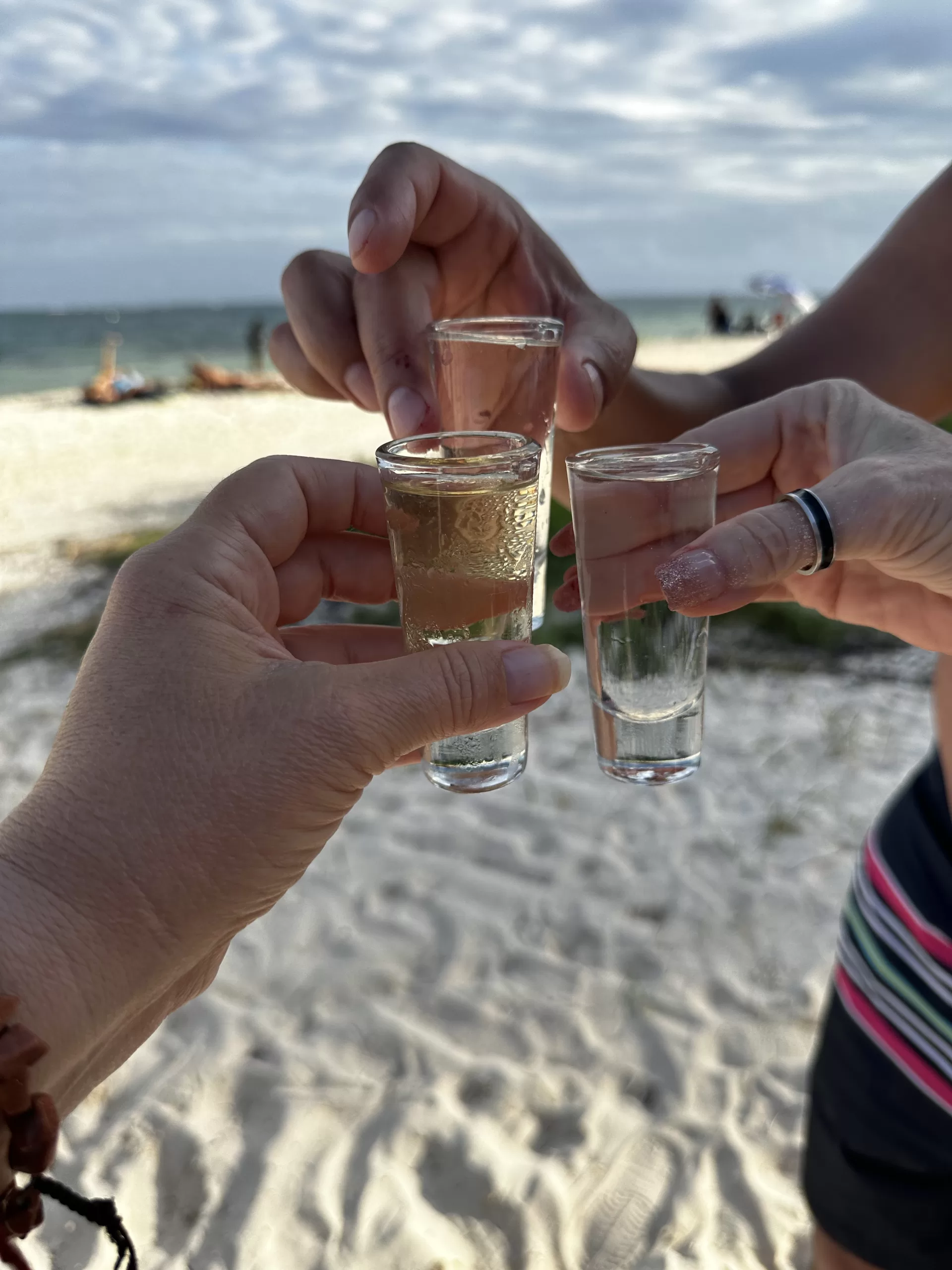 Three hands embrace shot glasses filled with golden tequila, capturing a moment of camaraderie and enjoyment against the backdrop of a sun-drenched beach.