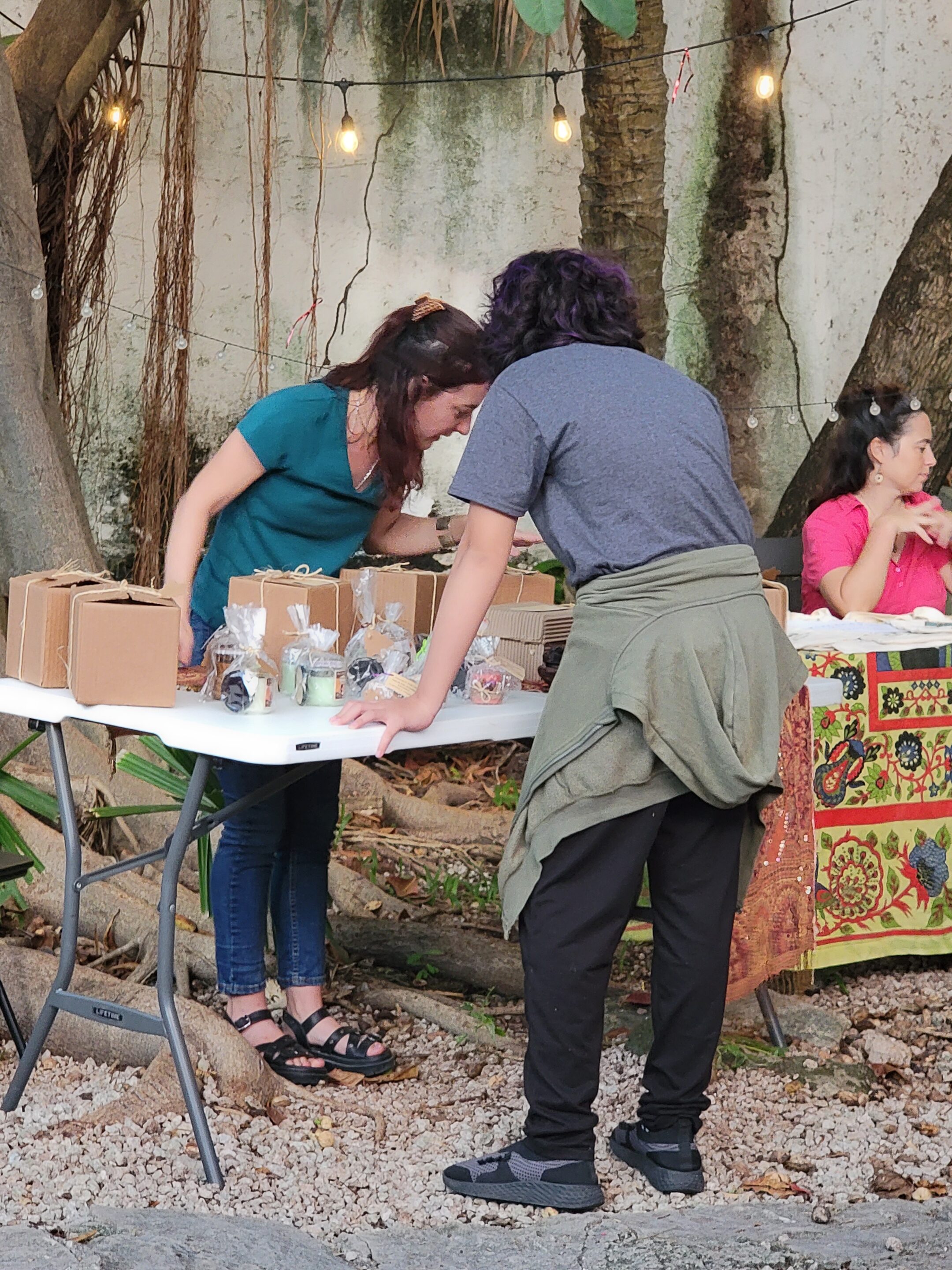 Two ladies standing outside at a table with several cardboard boxes, looking closely at something while they arrange items for sale at the Christmas Posada with El Telar and Experience Puerto Morelos.