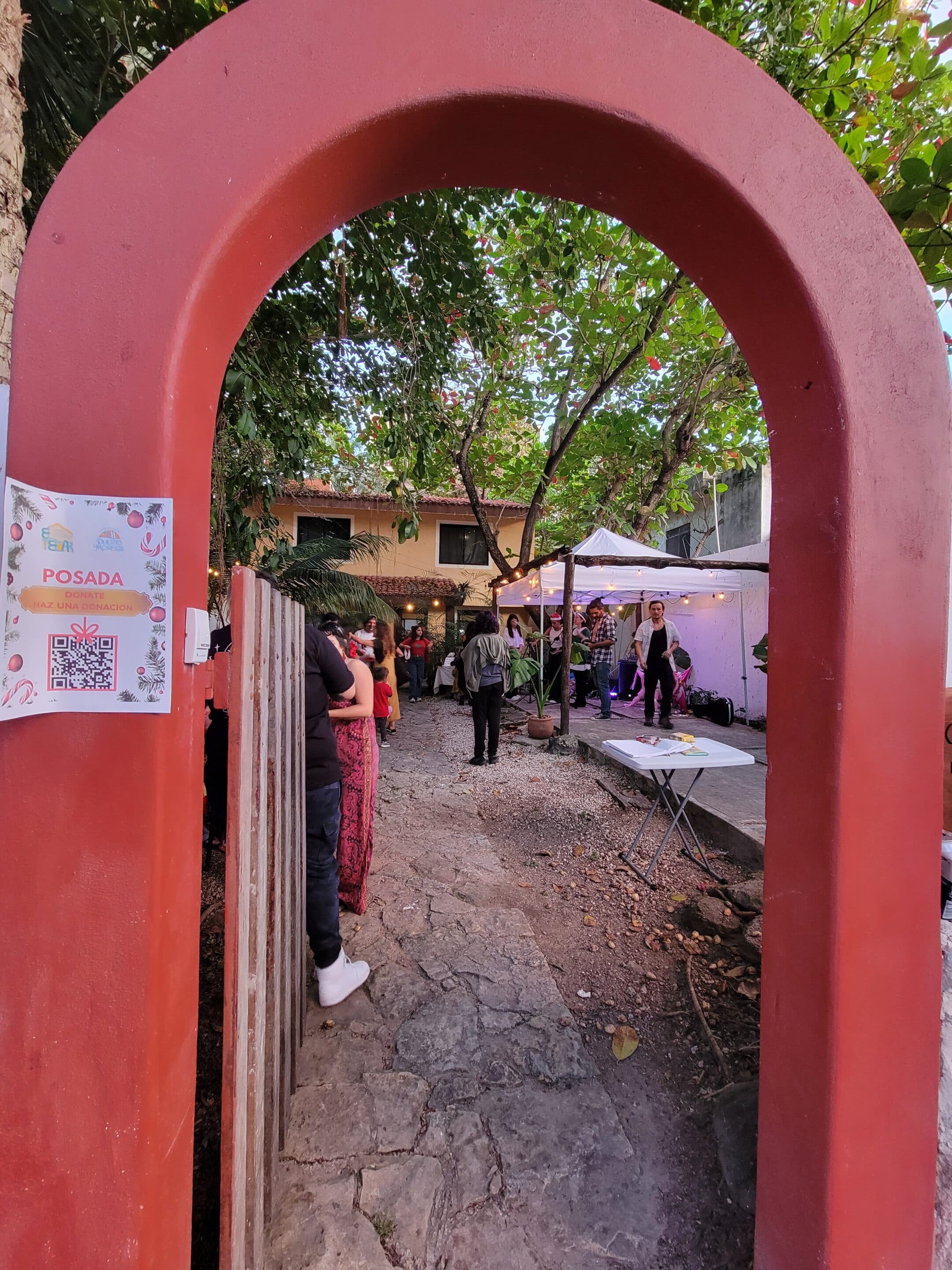 Photo of a a red archway with an open gate and a small sign stating "Posada". Beyond the archway, groups of people, a white tent, and a house are visible.