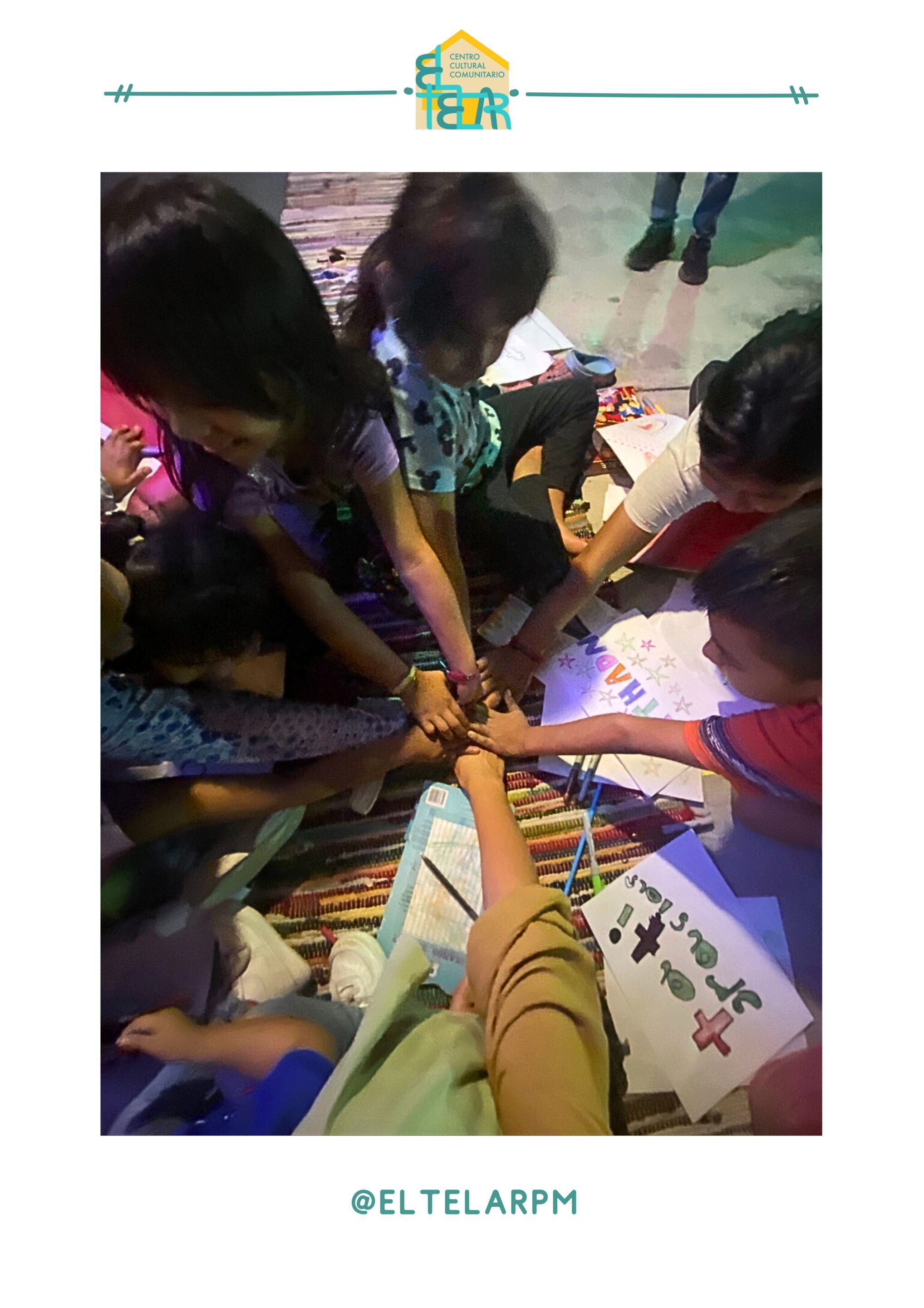 A group of children sitting in a circle on the floor at El Telar with their hands connected in the middle. Each child has a drawing they worked on during the class as well.