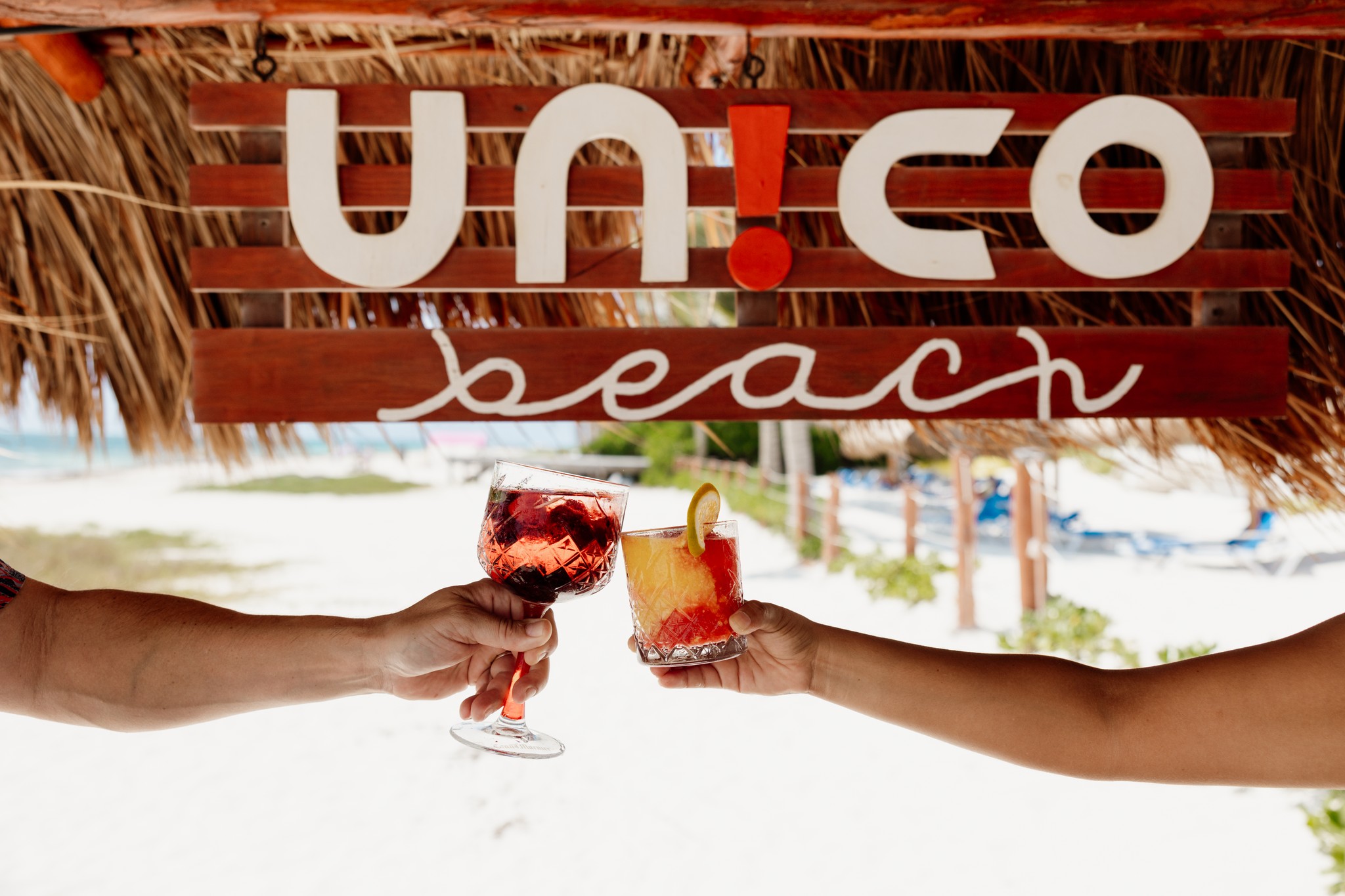 Two people holding cocktails in front of the Unico Beach sign.