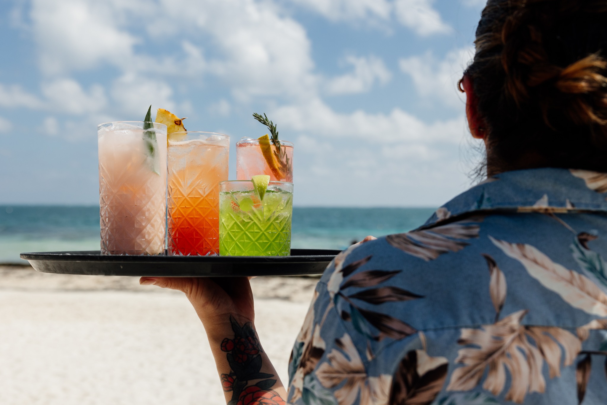 A waiter holds a serving tray with 4 colorful cocktails on it in front of the ocean at Unico Beach Club in Puerto Morelos.