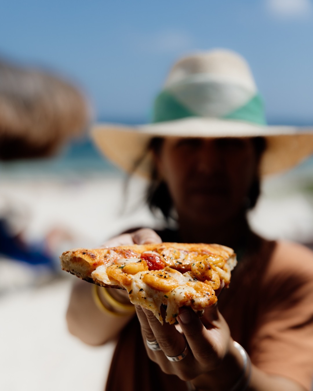 A woman stands on the beach at Unico Beach Club holding out a piece of pizza with cheese, shrimp and other veggies.
